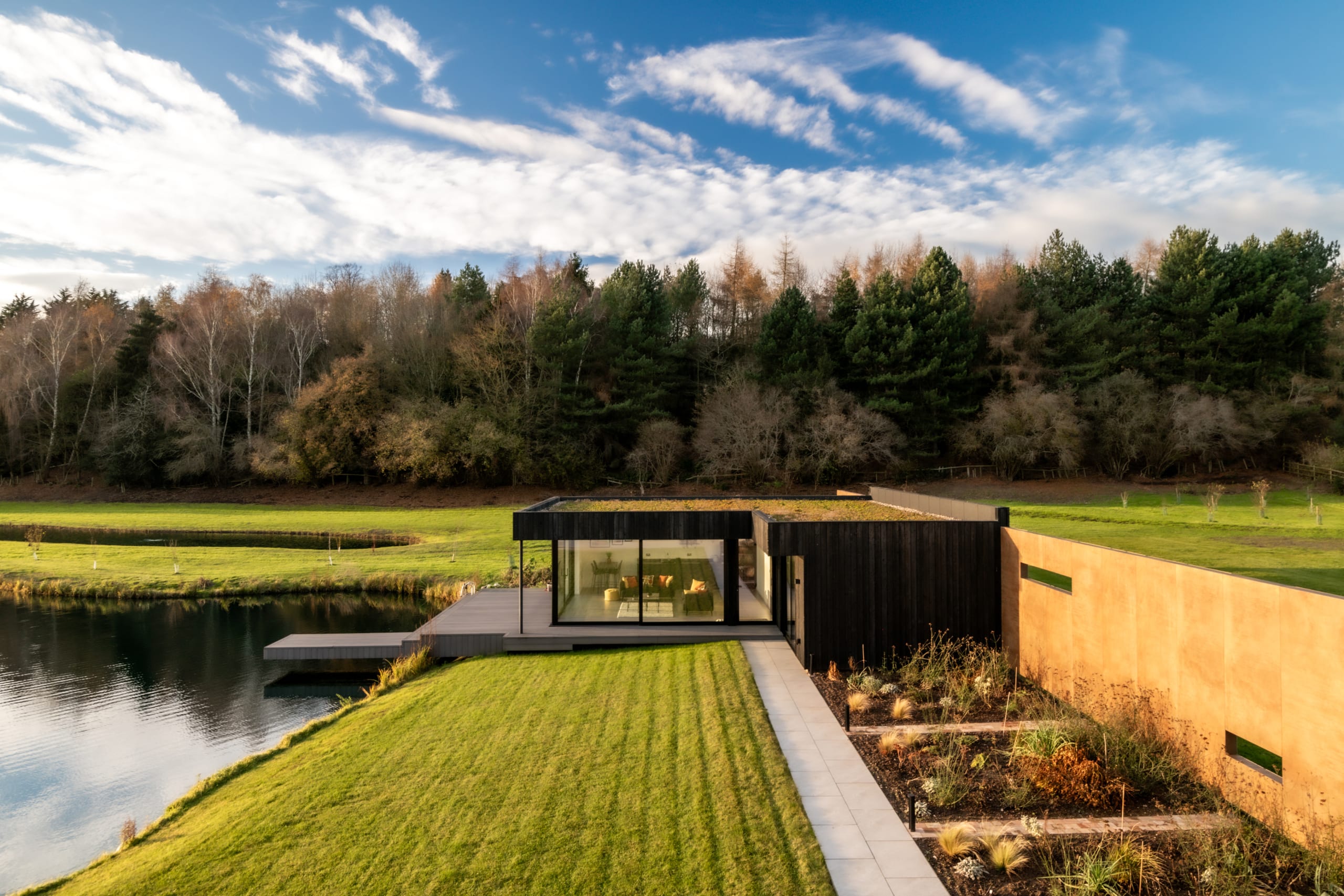 Modern single-story home with black timber cladding, green roof, and large glass windows overlooking a tranquil pond
